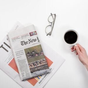 Two news papers on a white surface, with a pair of glasses and a hand holding a cup of coffee in a white mug, looking from the top down