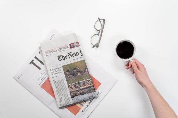 Two news papers on a white surface, with a pair of glasses and a hand holding a cup of coffee in a white mug, looking from the top down